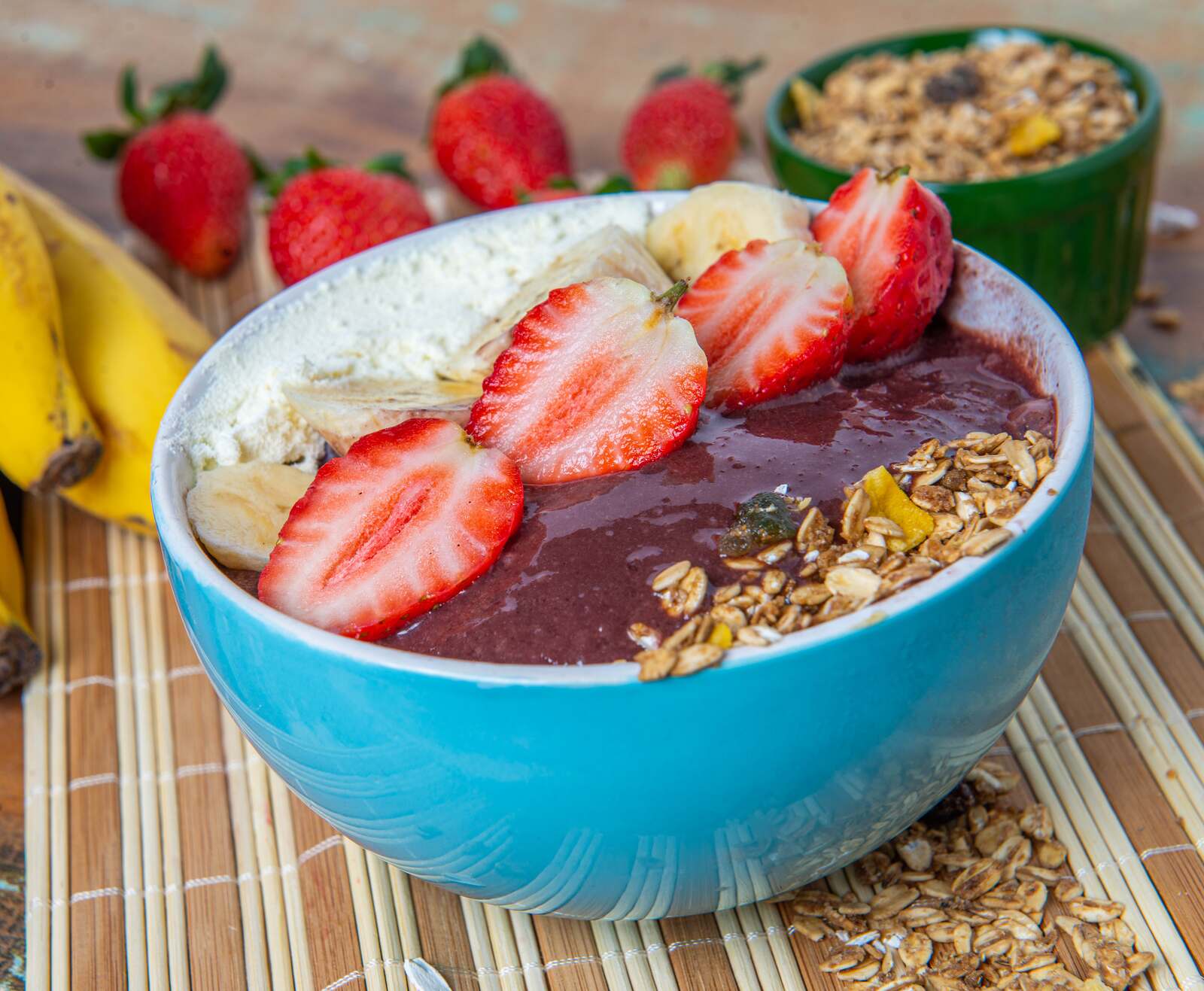 A blue bowl of açaí na tigela topped with sliced strawberries, banana, and granola, with bananas, strawberries, and a pot of granola around it