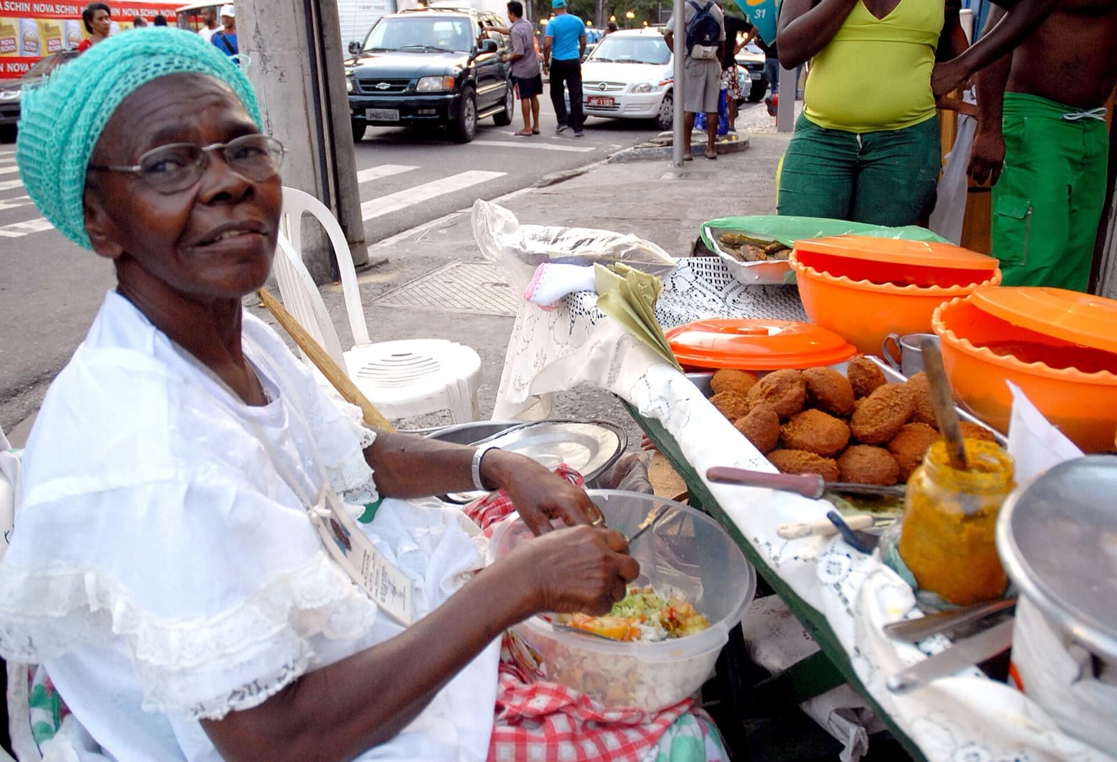 A baiana de acarajé in white lace preparing an acarajé at her street stand in Salvador, with fritters and pots of toppings on the table