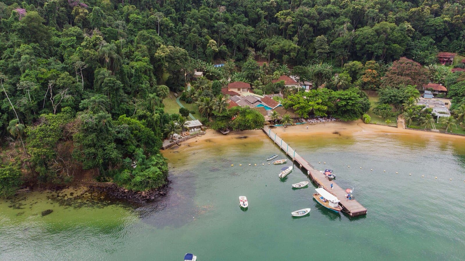Aerial view of a beach on Ilha da Gipóia in Angra dos Reis, with a pier, boats moored in green water, and rainforest behind