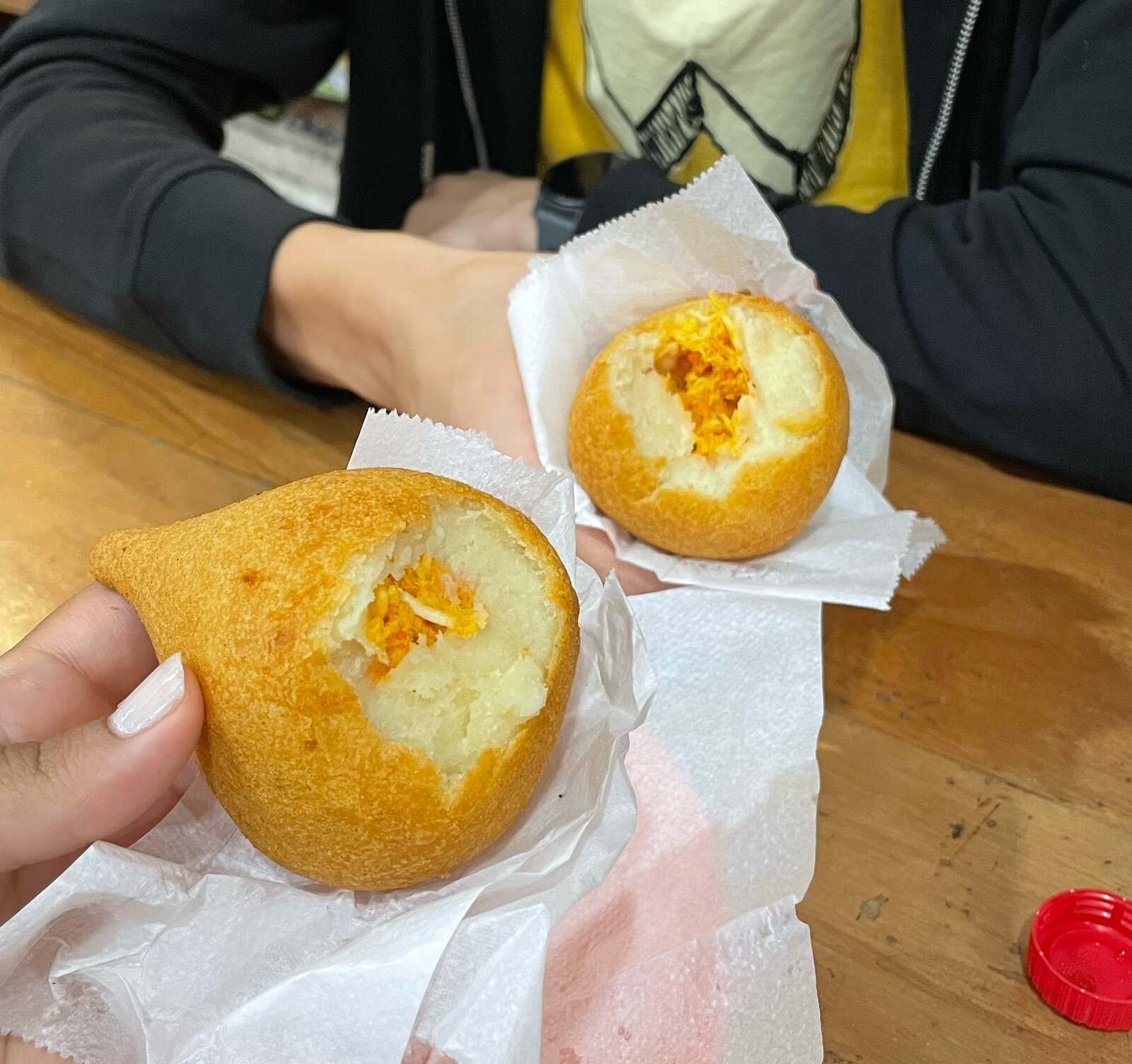 Two coxinhas bitten open to show the shredded chicken filling inside the fried dough, held in paper napkins over a wooden table