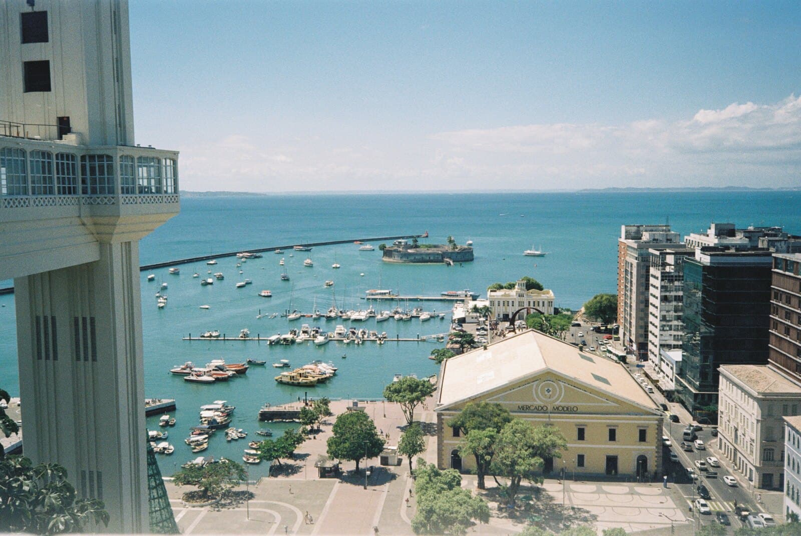The Elevador Lacerda's tower at the left and the view it opens over the Bay of All Saints, with boats moored off the marina, the round Forte de São Marcelo offshore, and the yellow Mercado Modelo below