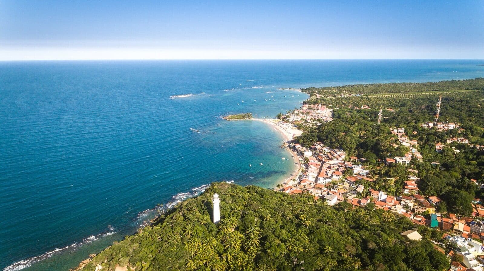 Aerial view of Morro de São Paulo on Tinharé island, with the white Farol do Morro lighthouse on the palm-covered headland above the village rooftops and the beaches curving away along the reef