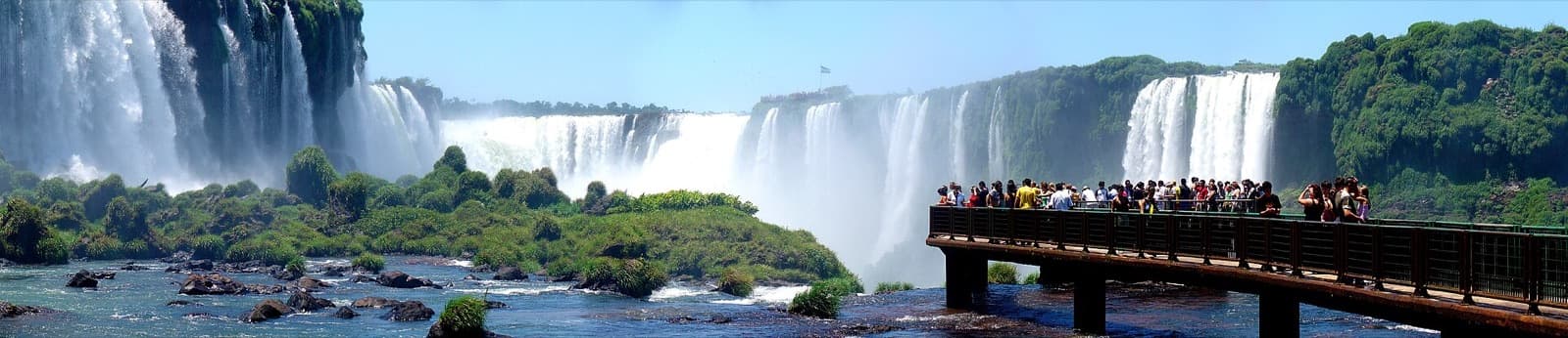 Wide panorama of Iguazu Falls with visitors crowding the Brazilian walkway that juts out toward the Garganta do Diabo