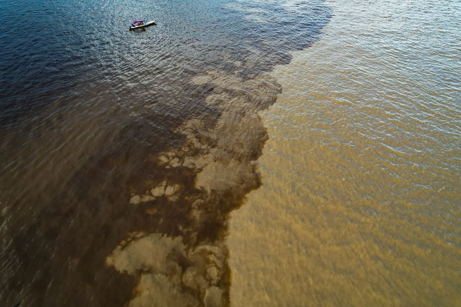 Aerial view of the Meeting of the Waters near Manaus, with the dark Rio Negro and the sandy Solimões River touching without mixing and a small boat on the color line