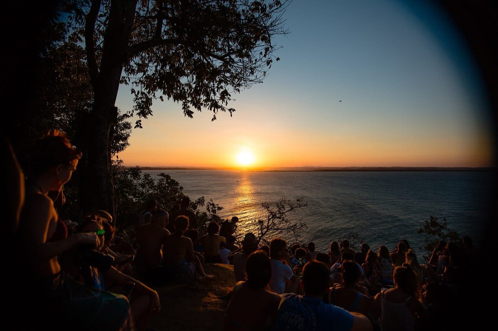 Crowd sitting on a hillside viewpoint in Morro de São Paulo at golden hour, watching the sun drop over the water toward the mainland