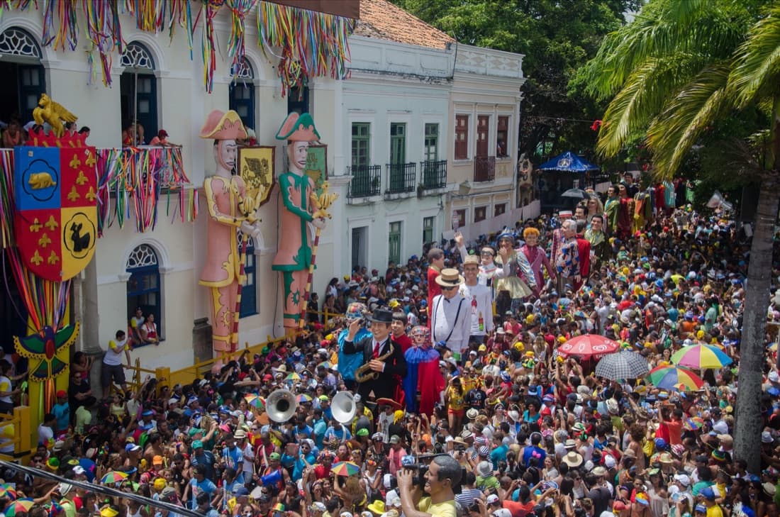 Two giant carnival puppets, the bonecos gigantes, towering over a packed crowd in front of colonial buildings during Carnival in Olinda, Pernambuco.