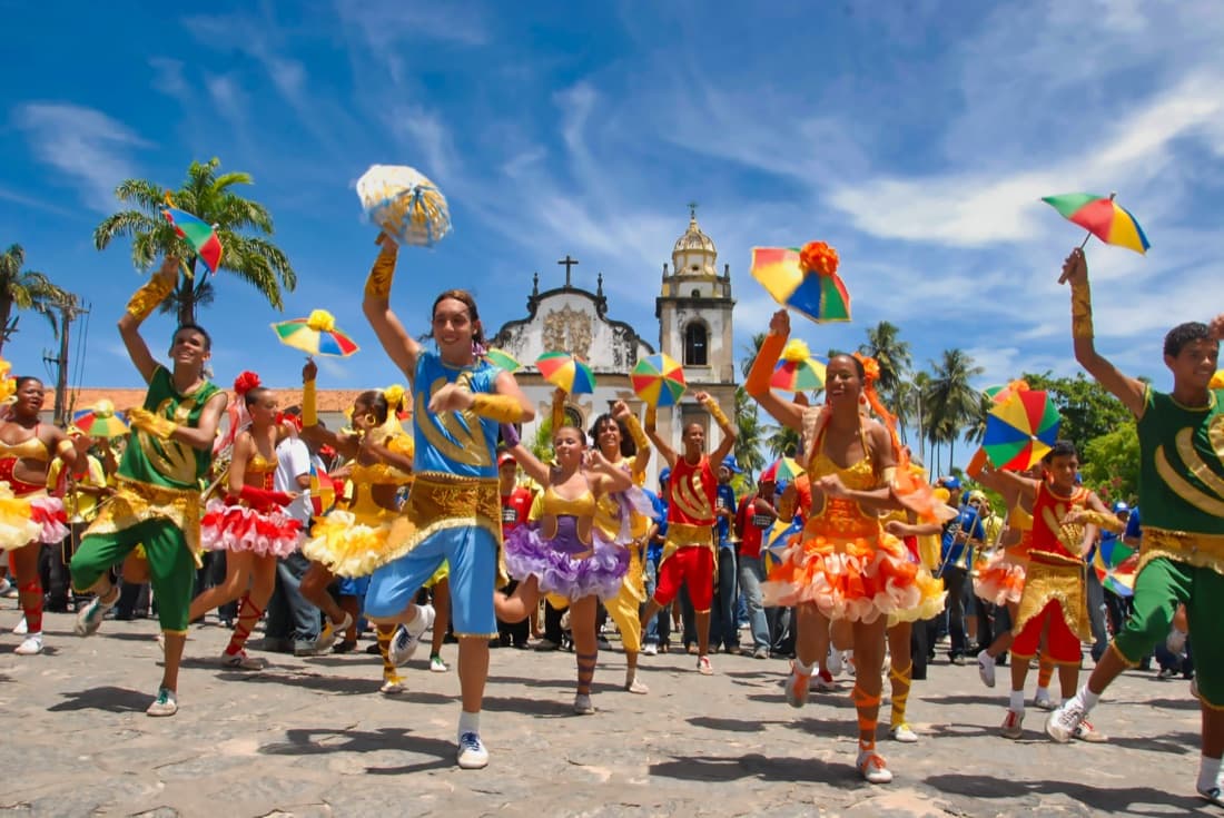 Frevo dancers in colorful costumes leaping with small umbrellas in front of a colonial church in Olinda, Pernambuco.