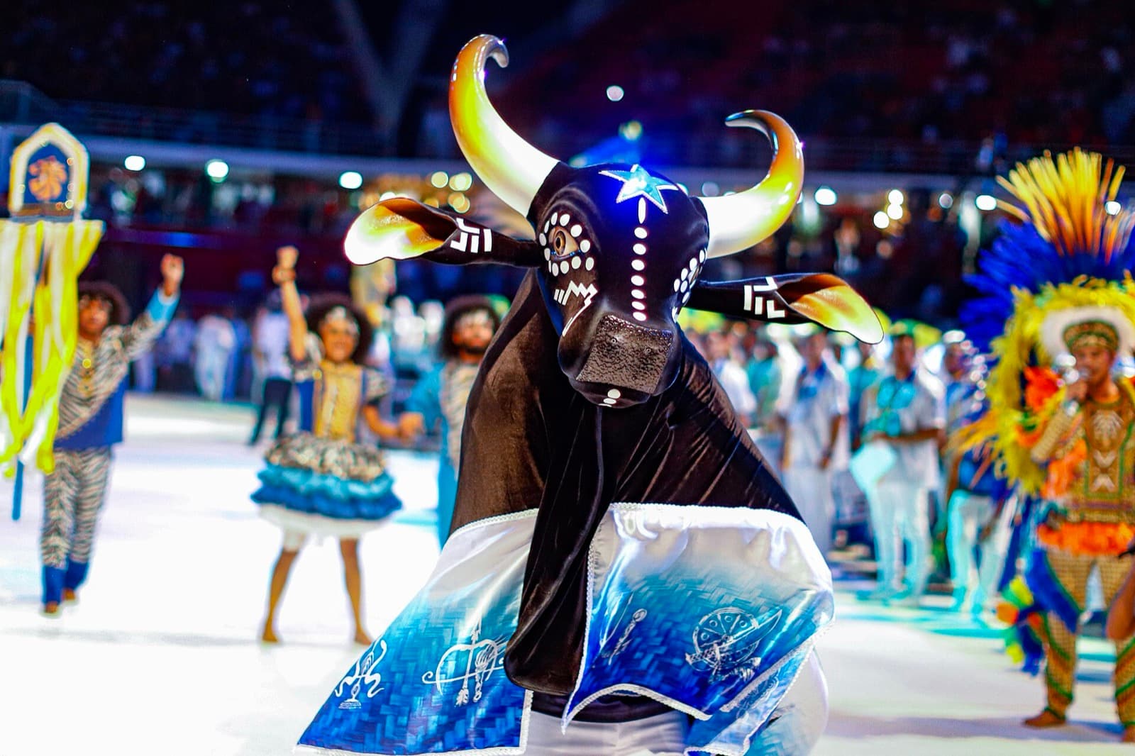 The Caprichoso ox, black with a blue star on its forehead and a blue and white skirt, performing under lights in the Bumbódromo arena at the Parintins festival.