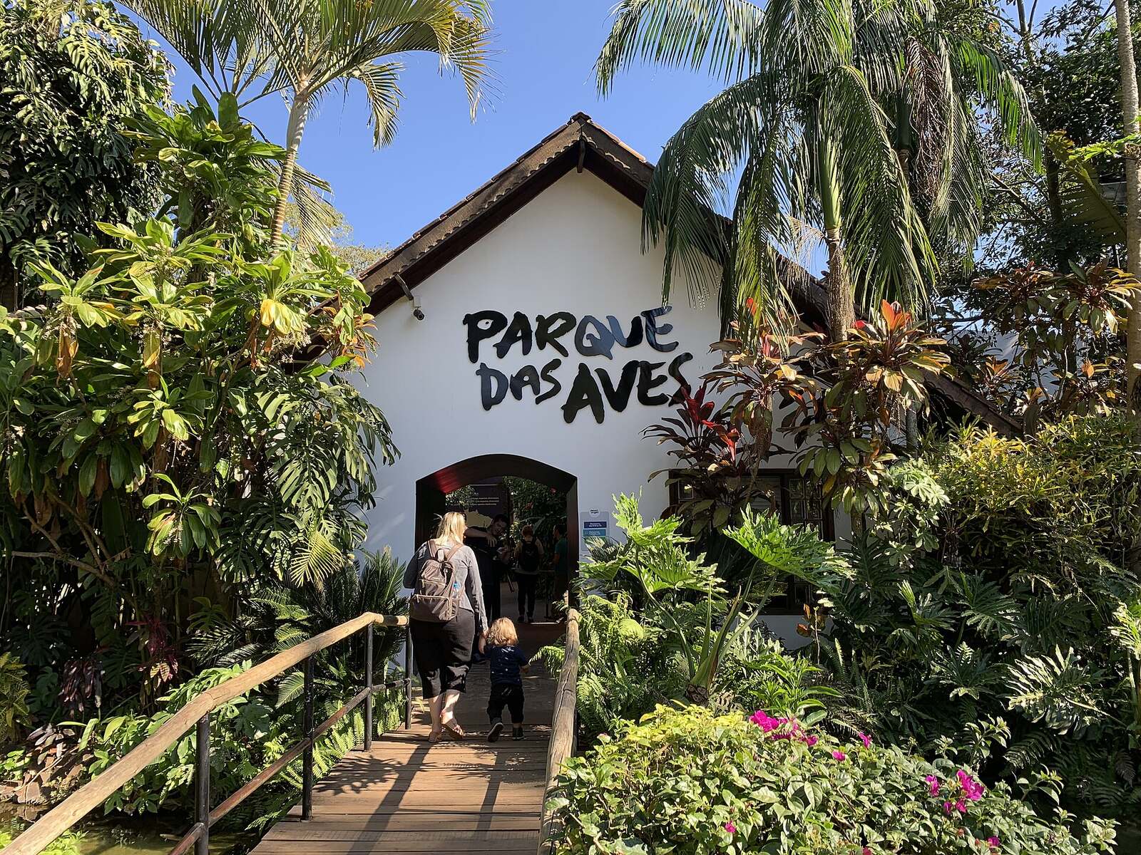 Visitors walking across a wooden bridge into the palm-shaded entrance of Parque das Aves in Foz do Iguaçu