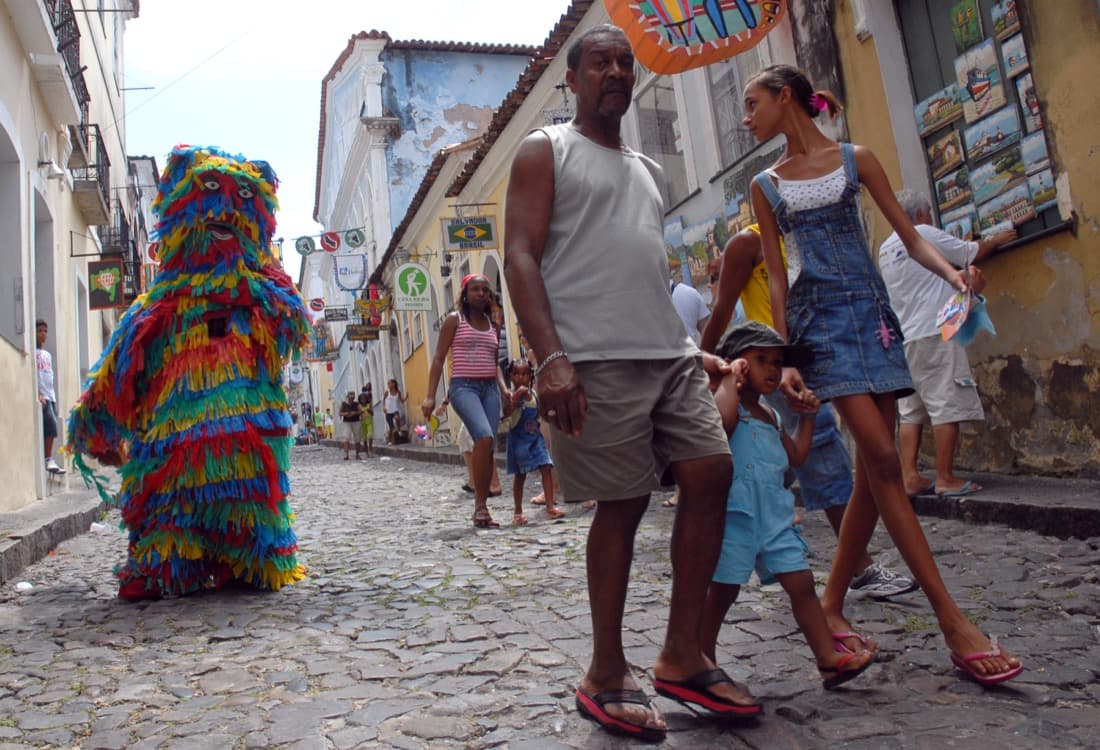 A person in a colorful fringed costume walking down a cobbled street in Pelourinho, Salvador's historic center, at the start of Carnival.