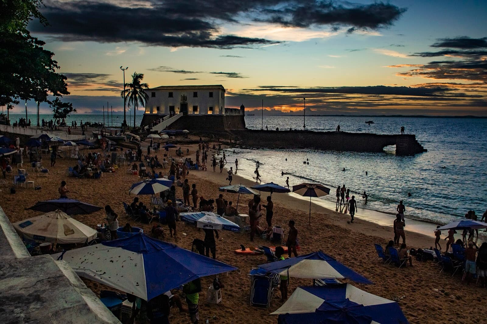 Beach umbrellas and swimmers at Porto da Barra beach at dusk, with the Santa Maria fort lit up in the background