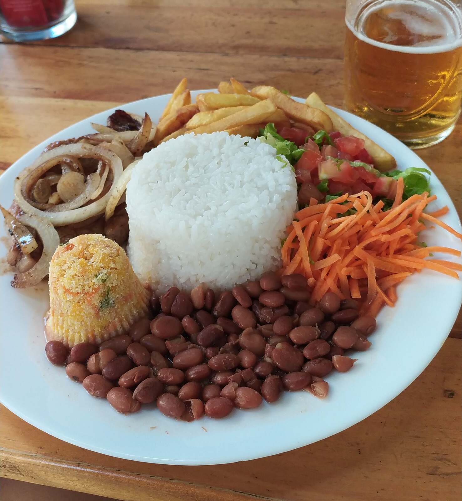 A prato feito on a wooden table: a molded dome of white rice with brown beans, fries, tomato and lettuce salad, shredded carrot, and a steak with grilled onions, with a glass of beer behind