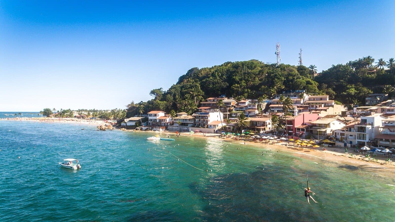 Aerial view of Primeira Praia in Morro de São Paulo, with guesthouses stacked up the wooded hillside, yellow umbrellas along the small strip of sand, boats moored offshore, and a zipline rider dropping toward the water