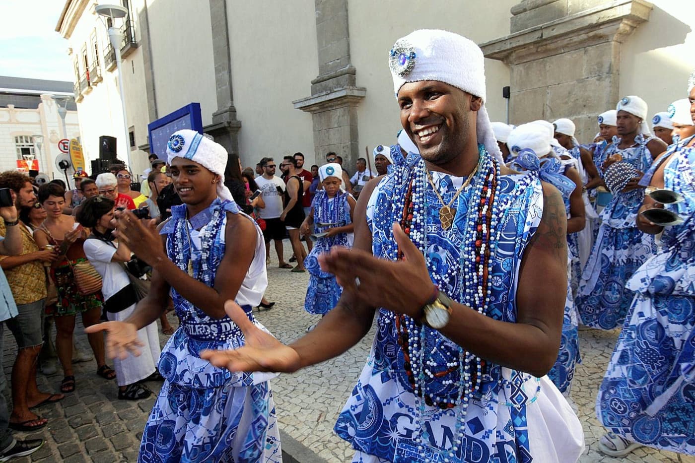 Performers in matching blue and white costumes and strings of beads dancing through a street crowd during Carnival in Salvador, Bahia.