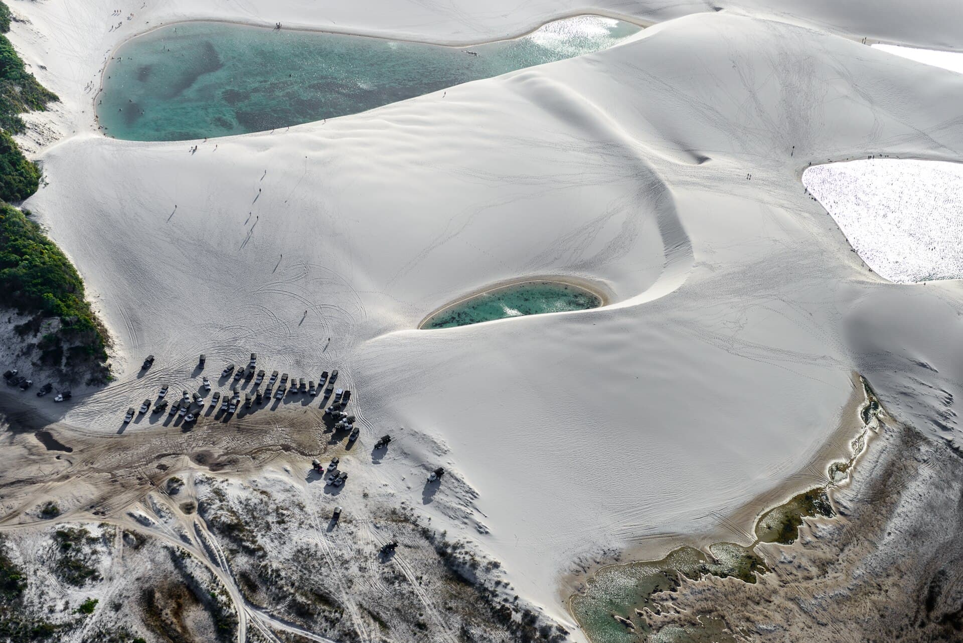 Aerial view of a row of tour trucks parked on the sand where the vegetation ends and the white dunes and turquoise lagoons of Lençóis Maranhenses begin