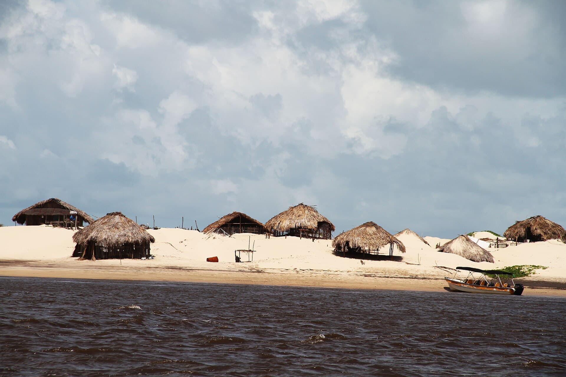 Thatched huts and a moored tour boat on the sandbar at Caburé, seen from the Rio Preguiças under a cloudy sky