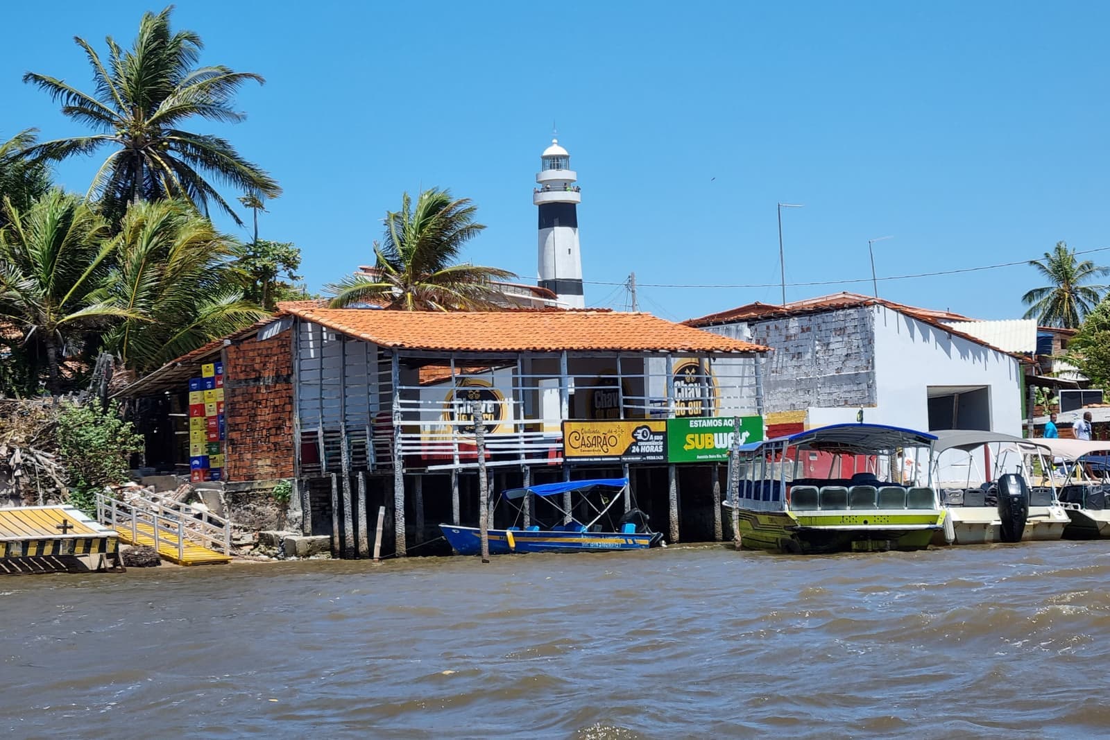 The black-and-white striped Farol Preguiças lighthouse rising behind palm trees and a riverside building on stilts at Mandacaru, with tour boats moored along the Rio Preguiças