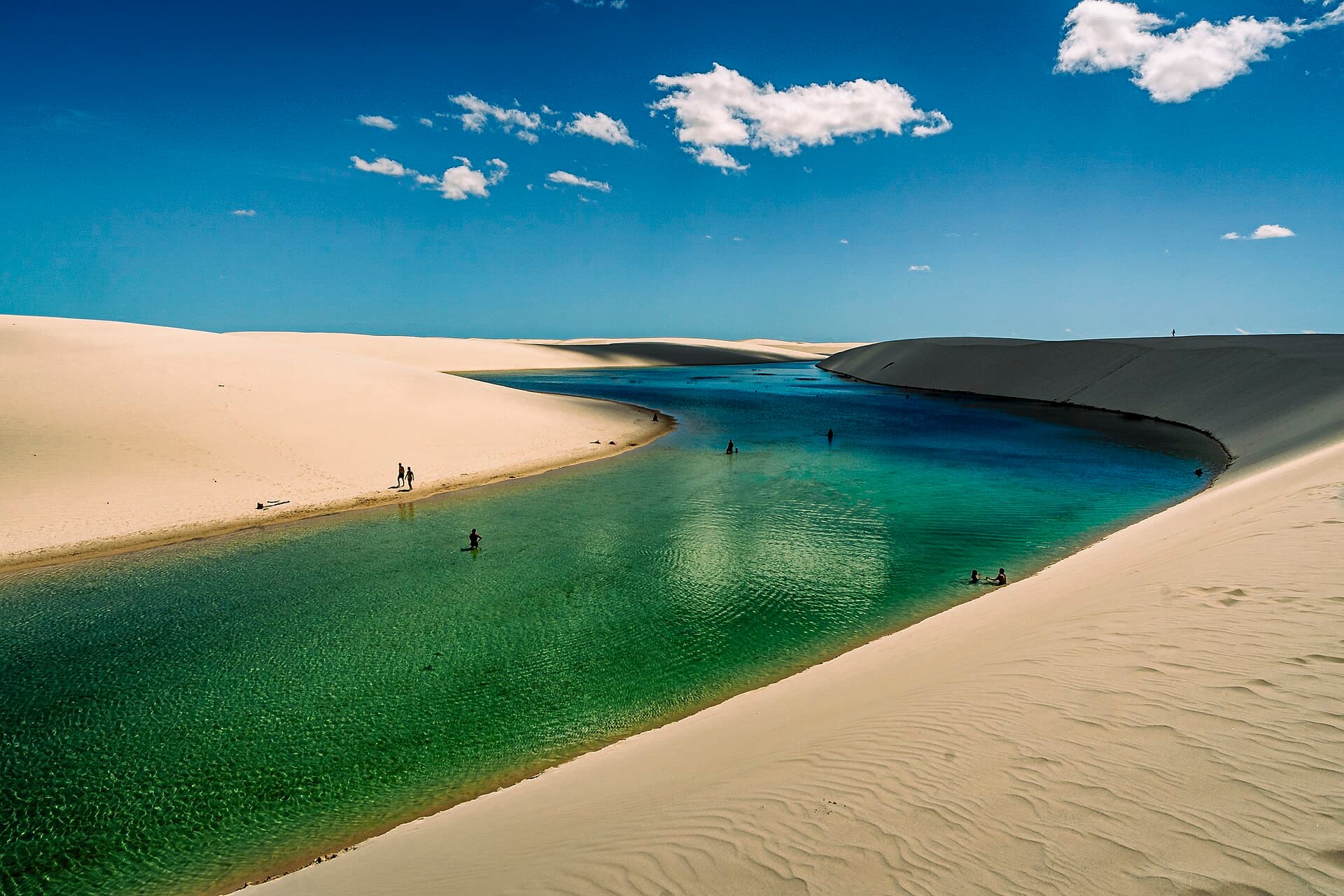 Swimmers wading in a long emerald lagoon between white sand dunes on the Lagoa Bonita circuit in Lençóis Maranhenses National Park, under a blue sky with scattered clouds