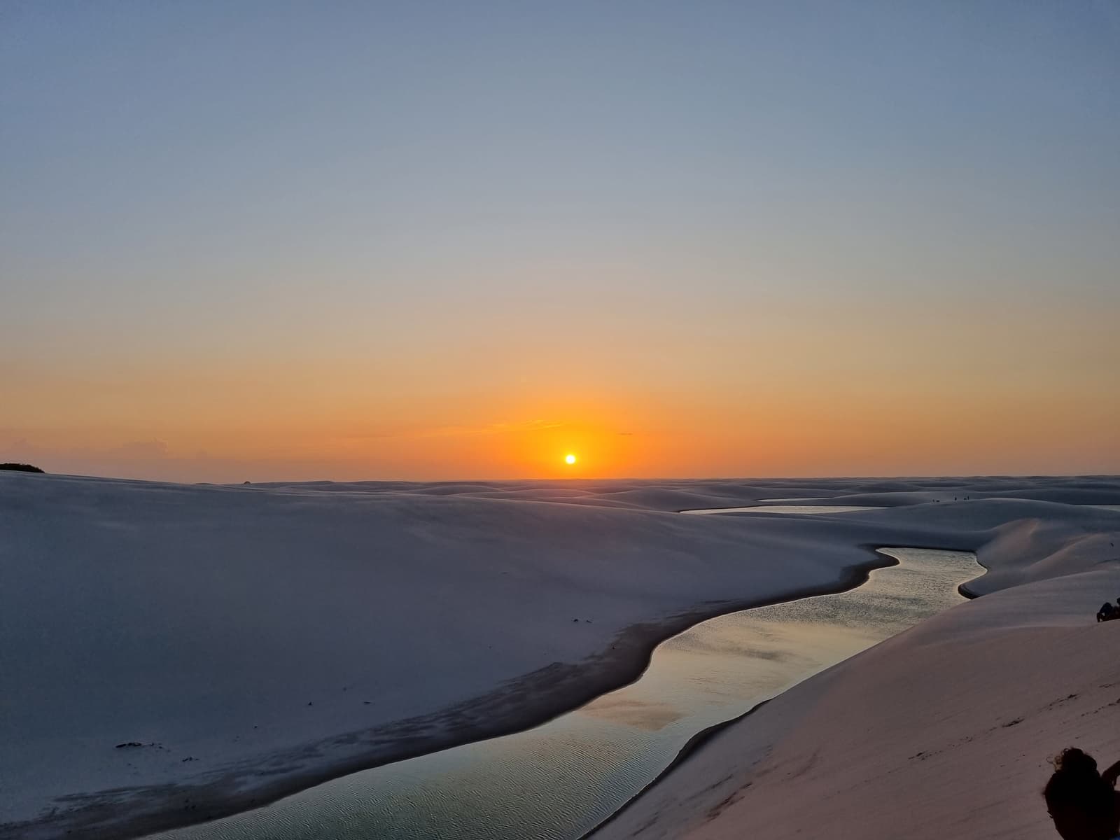 The sun setting over the white dunes of Lençóis Maranhenses, with a narrow lagoon reflecting the orange sky between the dunes below