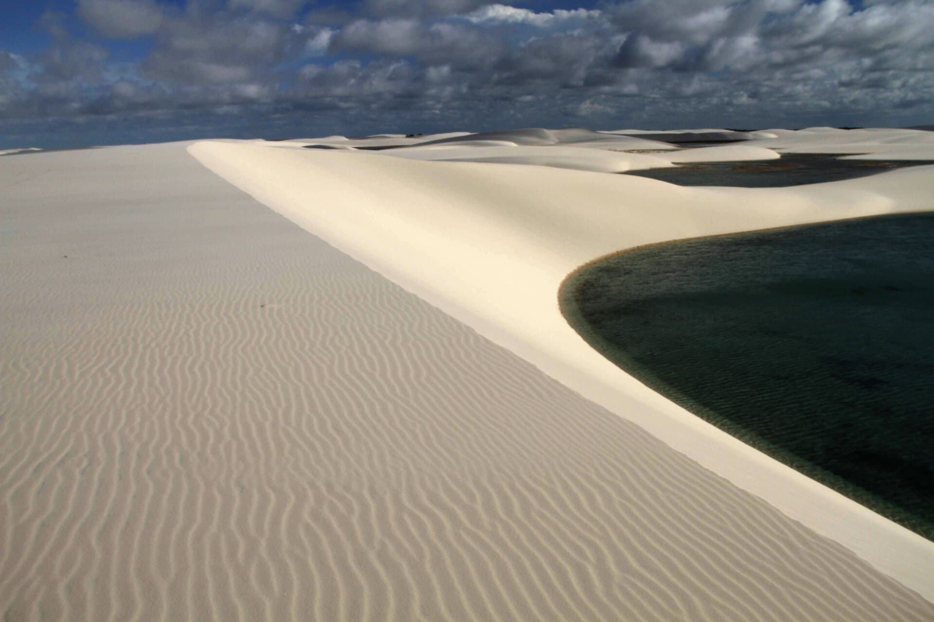 A knife-edged white dune curving around the dark blue water of the Lagoa da Gaivota near Santo Amaro do Maranhão, with more dunes rolling to the horizon under a cloudy sky