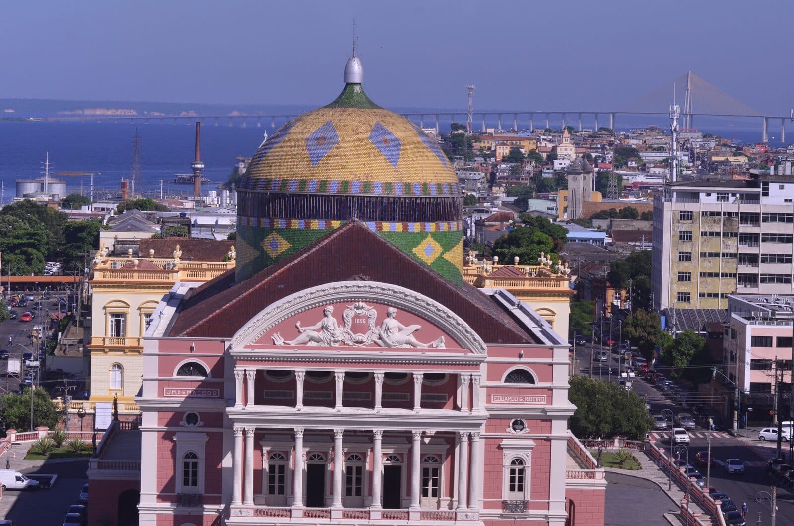 The Teatro Amazonas opera house in Manaus seen from above, its pink and white facade below a dome tiled in green, yellow and blue, with the city, the Rio Negro, and a cable-stayed bridge behind it