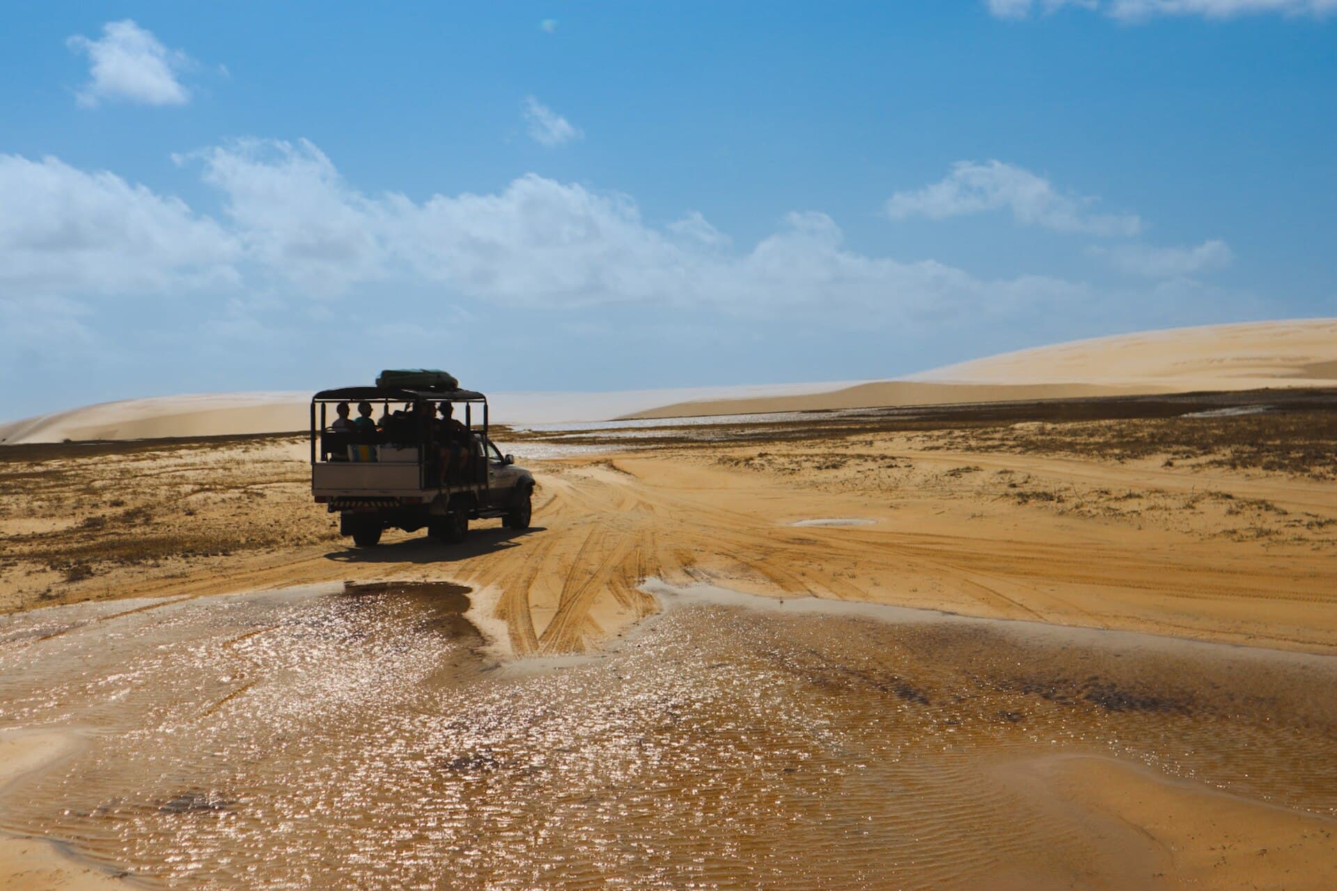 An open-backed Toyota 4x4 tour truck splashing through a flooded sand track toward the white dunes of Lençóis Maranhenses National Park
