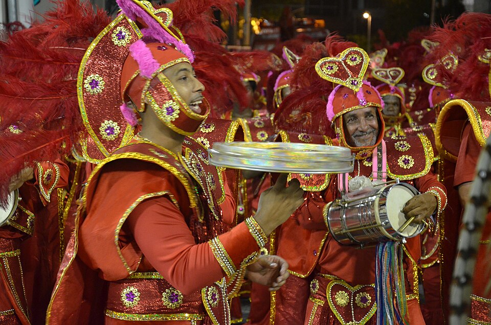 Carnival parade performers in red costumes playing percussion during a samba procession in Rio de Janeiro.