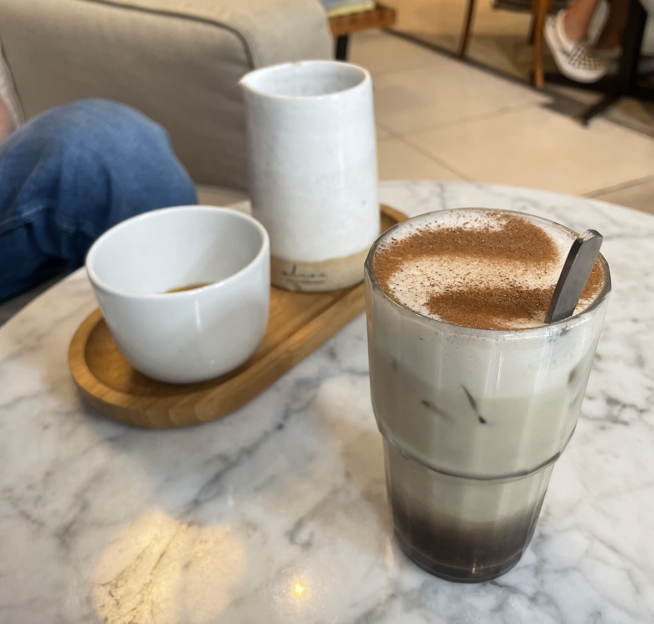 An iced latte dusted with cinnamon, an espresso, and a serving carafe on a marble table at Elisa Café in Belo Horizonte